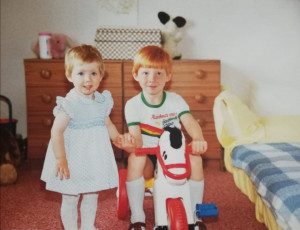 Claire and Brian are small children holding hands in a bedroom, Brian is sitting on a rocking horse and they are both smiling